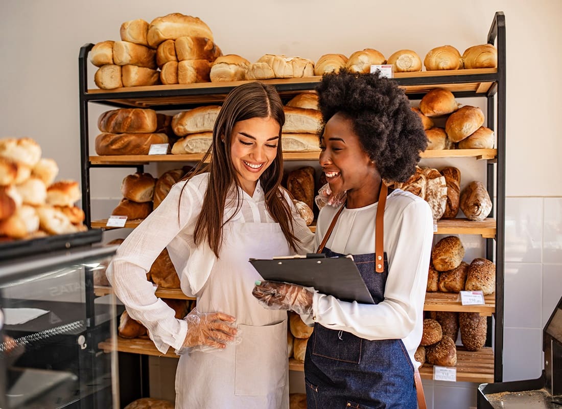 Read Our Reviews - Portrait of Two Cheerful Young Women Standing in Front of a Rack of Fresh Baked Bread While Looking at a Clipboard